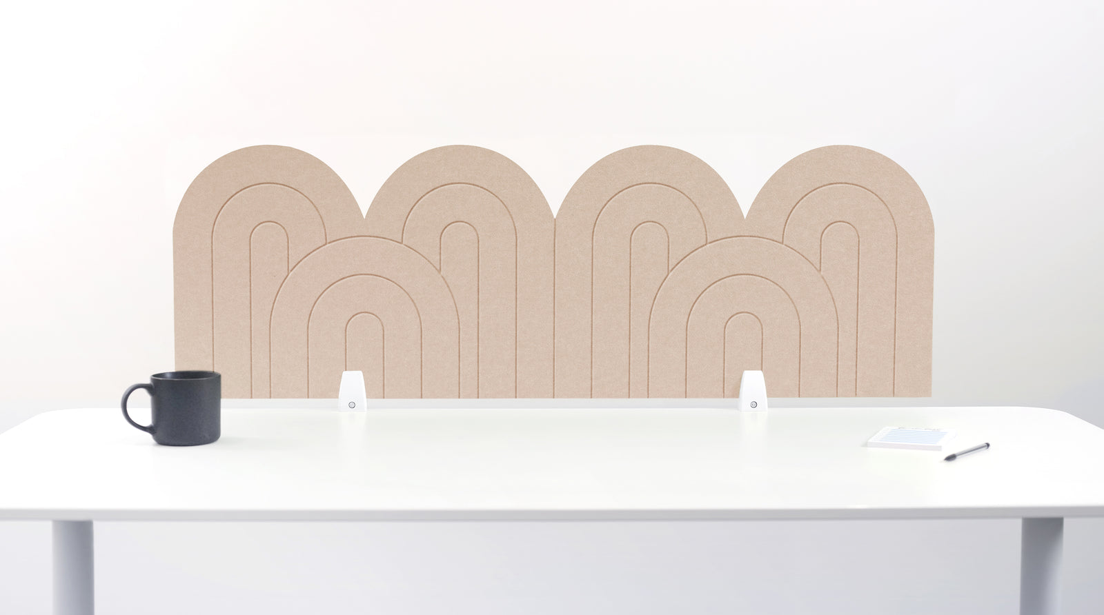 A light tan felt desk divider with a wavy, layered arch pattern, shown standing behind a white desk with a black mug on the left and a notepad on the right.