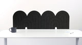 An dark gray felt desk divider with a wavy, layered arch pattern, shown standing behind a white desk with a black mug on the left and a notepad on the right.