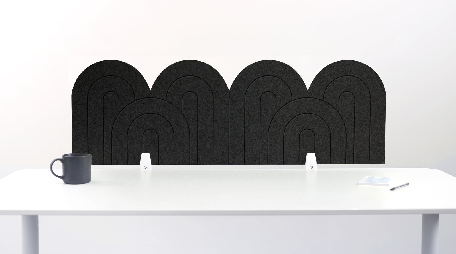 An dark gray felt desk divider with a wavy, layered arch pattern, shown standing behind a white desk with a black mug on the left and a notepad on the right.