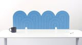 An light blue felt desk divider with a wavy, layered arch pattern, shown standing behind a white desk with a black mug on the left and a notepad on the right.