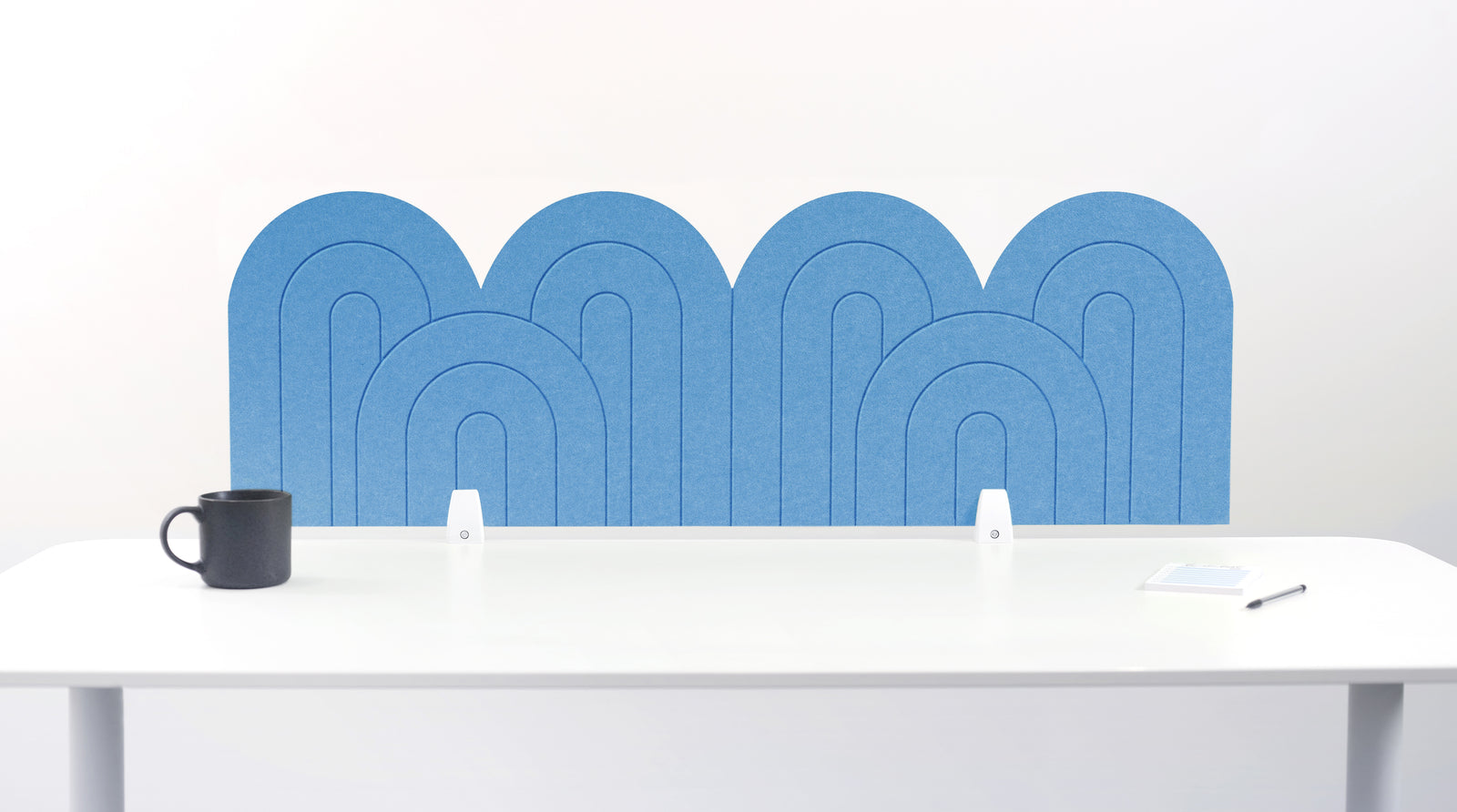 An light blue felt desk divider with a wavy, layered arch pattern, shown standing behind a white desk with a black mug on the left and a notepad on the right.