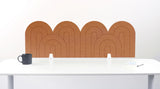 A cinnamon colored felt desk divider with a wavy, layered arch pattern, shown standing behind a white desk with a black mug on the left and a notepad on the right.
