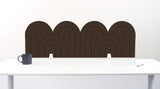 A brown felt desk divider with a wavy, layered arch pattern, shown standing behind a white desk with a black mug on the left and a notepad on the right.