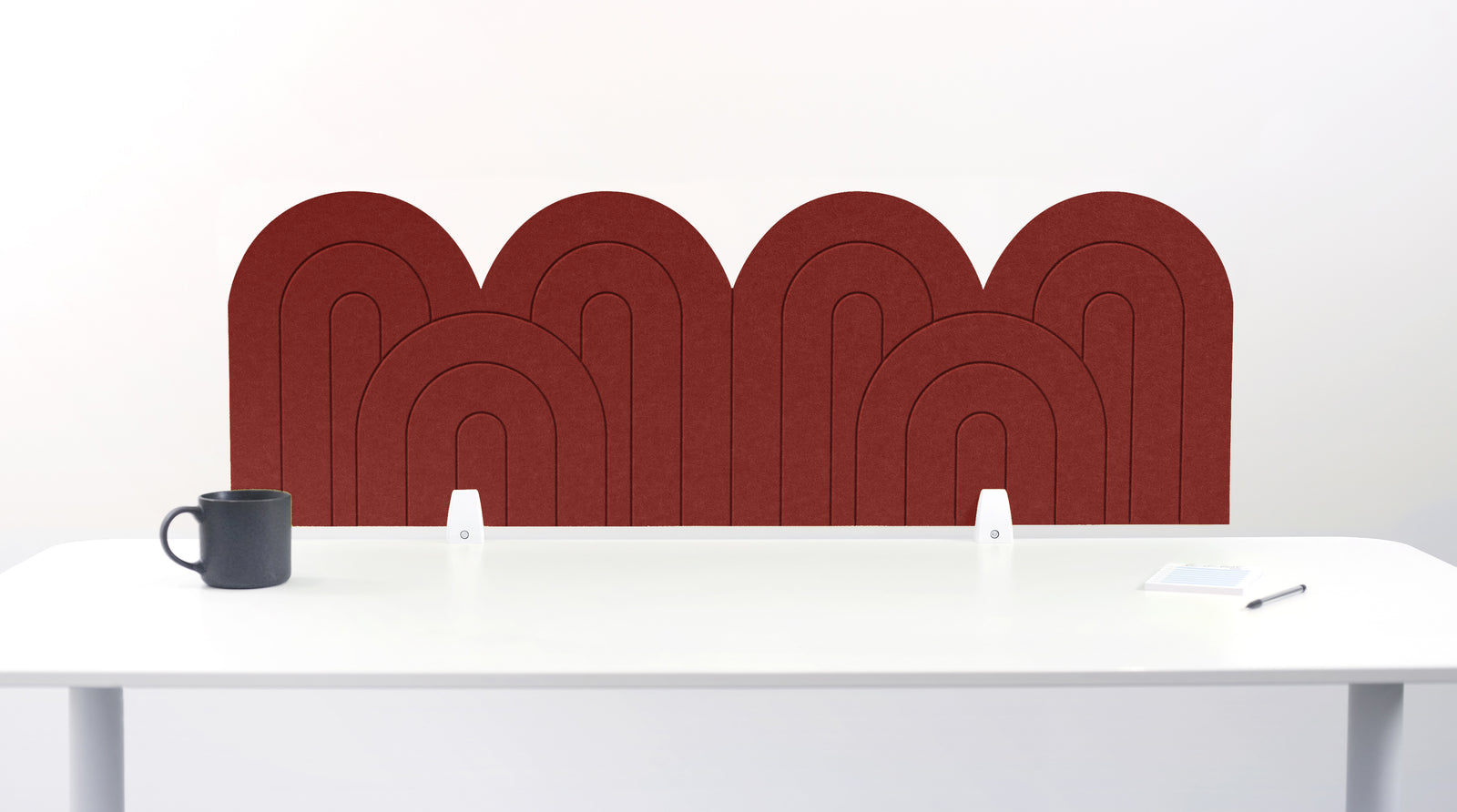 A crimson red felt desk divider with a wavy, layered arch pattern, shown standing behind a white desk with a black mug on the left and a notepad on the right.