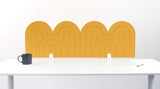 A yellow felt desk divider with a wavy, layered arch pattern, shown standing behind a white desk with a black mug on the left and a notepad on the right.