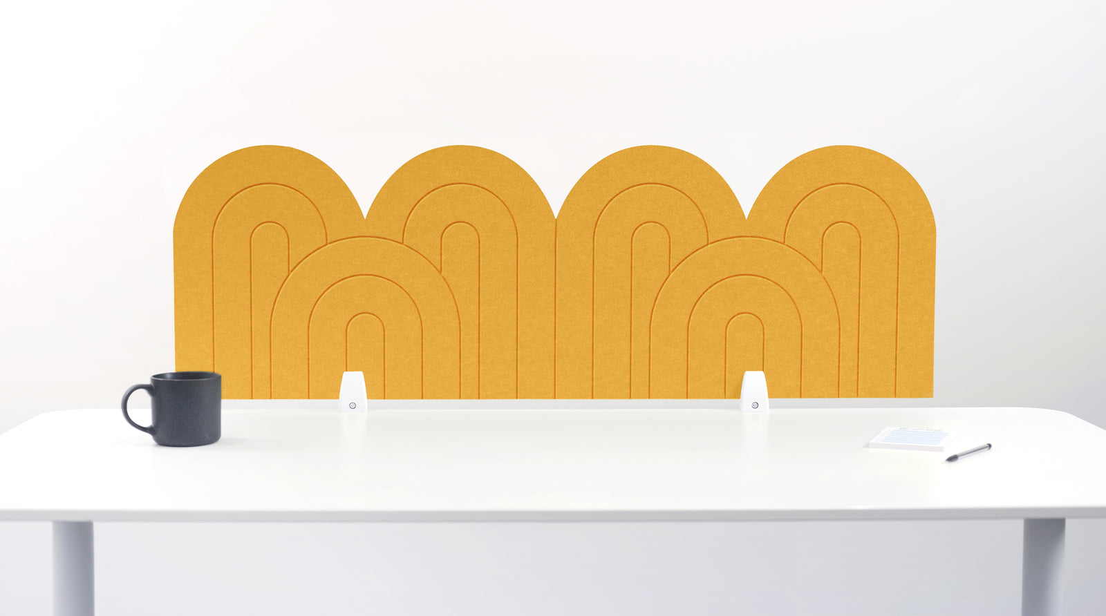 A yellow felt desk divider with a wavy, layered arch pattern, shown standing behind a white desk with a black mug on the left and a notepad on the right.