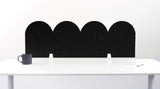 A black felt desk divider with a wavy, layered arch pattern, shown standing behind a white desk with a black mug on the left and a notepad on the right.