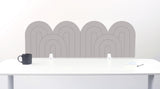 A fog colored felt desk divider with a wavy, layered arch pattern, shown standing behind a white desk with a black mug on the left and a notepad on the right.