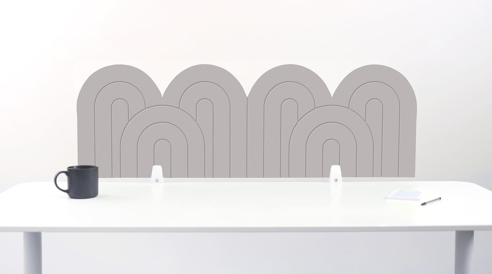 A fog colored felt desk divider with a wavy, layered arch pattern, shown standing behind a white desk with a black mug on the left and a notepad on the right.