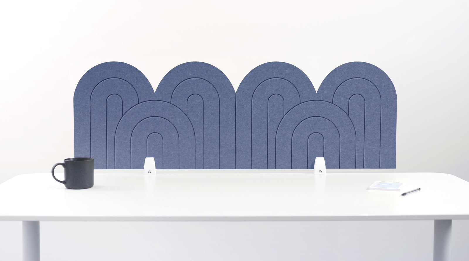 A blue indigo felt desk divider with a wavy, layered arch pattern, shown standing behind a white desk with a black mug on the left and a notepad on the right.