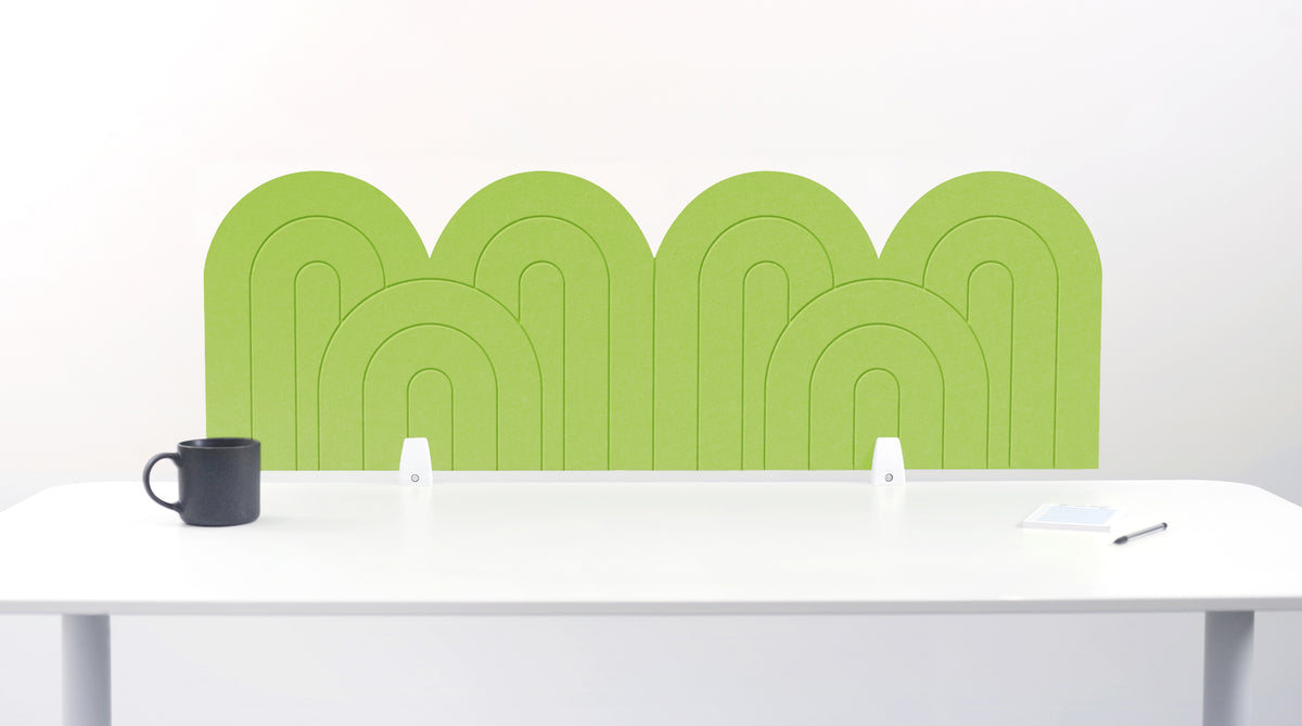 An lime green felt desk divider with a wavy, layered arch pattern, shown standing behind a white desk with a black mug on the left and a notepad on the right.