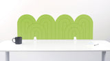 An lime green felt desk divider with a wavy, layered arch pattern, shown standing behind a white desk with a black mug on the left and a notepad on the right.