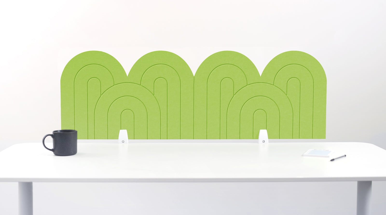An lime green felt desk divider with a wavy, layered arch pattern, shown standing behind a white desk with a black mug on the left and a notepad on the right.