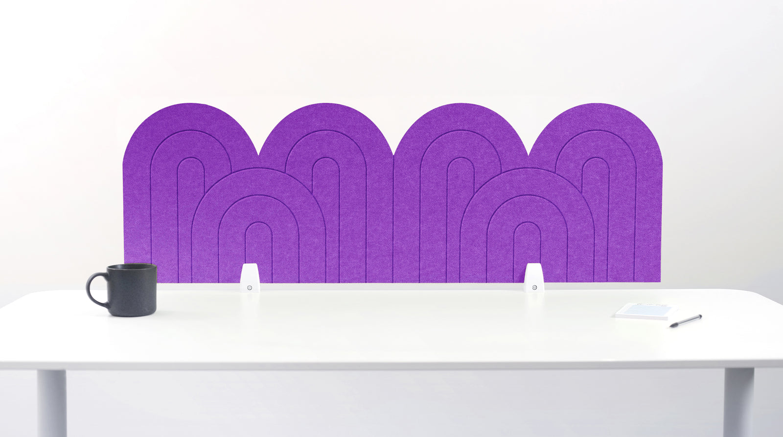 An purple felt desk divider with a wavy, layered arch pattern, shown standing behind a white desk with a black mug on the left and a notepad on the right.