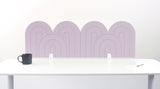 A light pink felt desk divider with a wavy, layered arch pattern, shown standing behind a white desk with a black mug on the left and a notepad on the right.