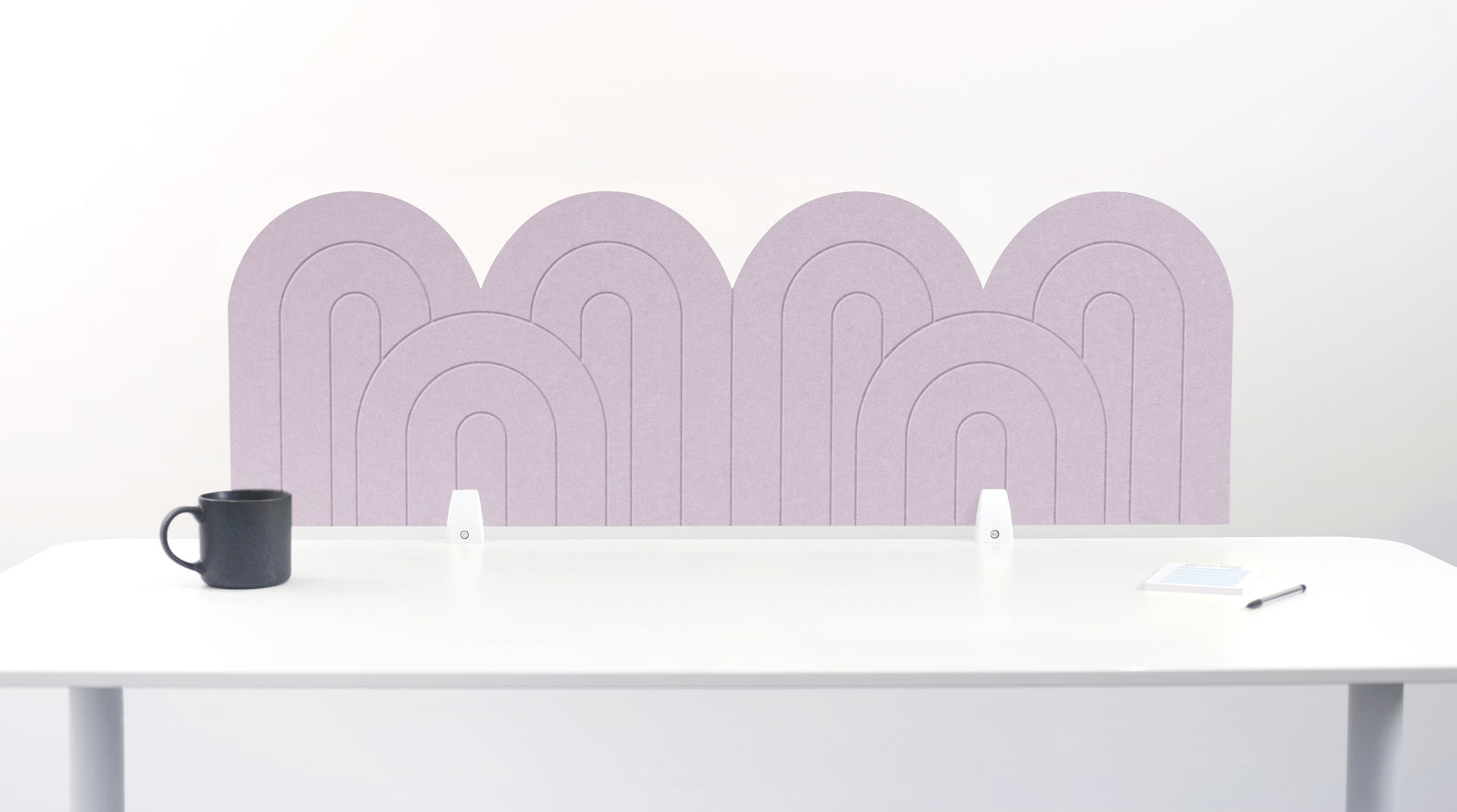 A light pink felt desk divider with a wavy, layered arch pattern, shown standing behind a white desk with a black mug on the left and a notepad on the right.