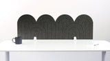 A light gray felt desk divider with a wavy, layered arch pattern, shown standing behind a white desk with a black mug on the left and a notepad on the right.