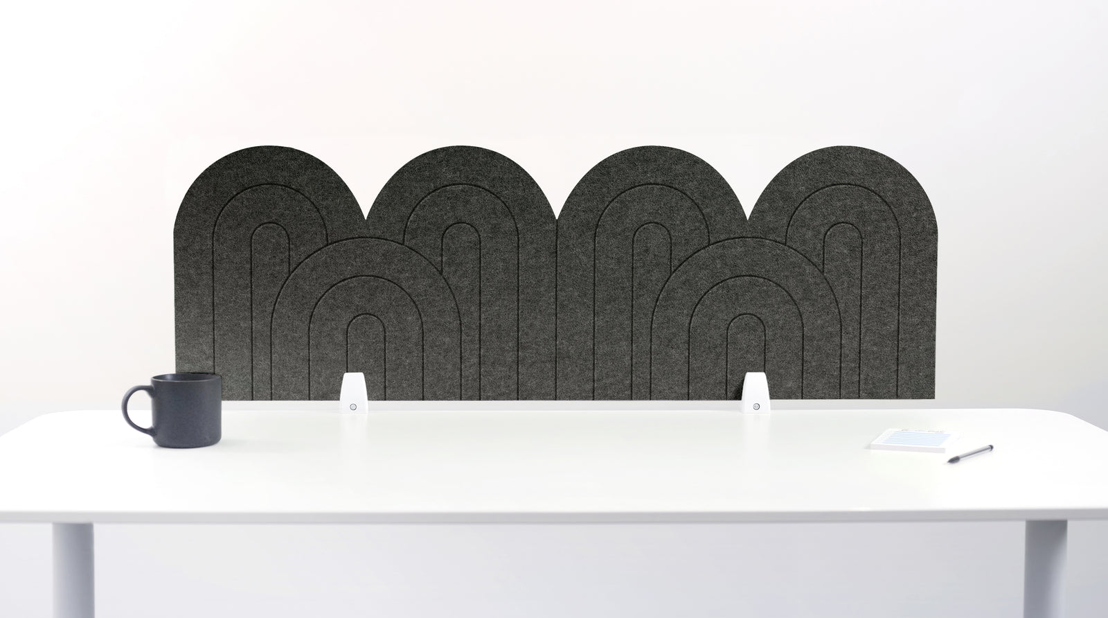 A light gray felt desk divider with a wavy, layered arch pattern, shown standing behind a white desk with a black mug on the left and a notepad on the right.