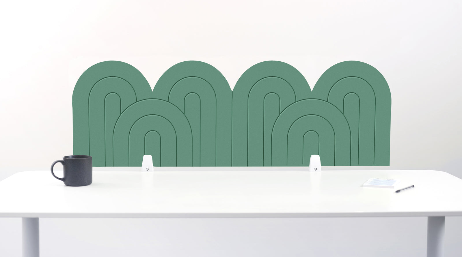A mint felt desk divider with a wavy, layered arch pattern, shown standing behind a white desk with a black mug on the left and a notepad on the right.