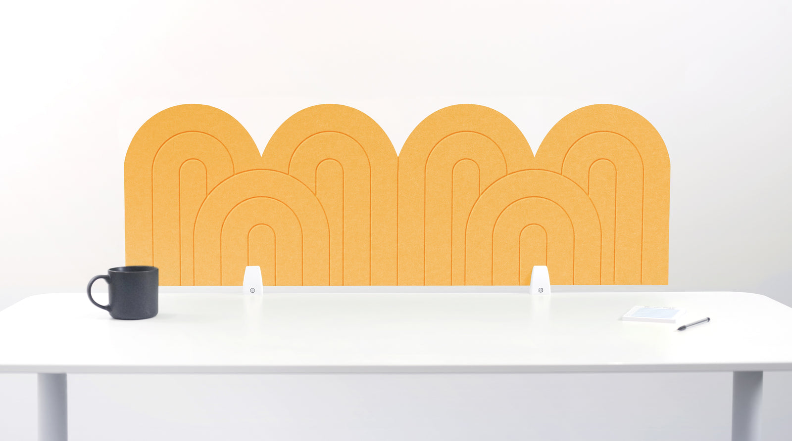 A yellow felt desk divider with a wavy, layered arch pattern, shown standing behind a white desk with a black mug on the left and a notepad on the right.