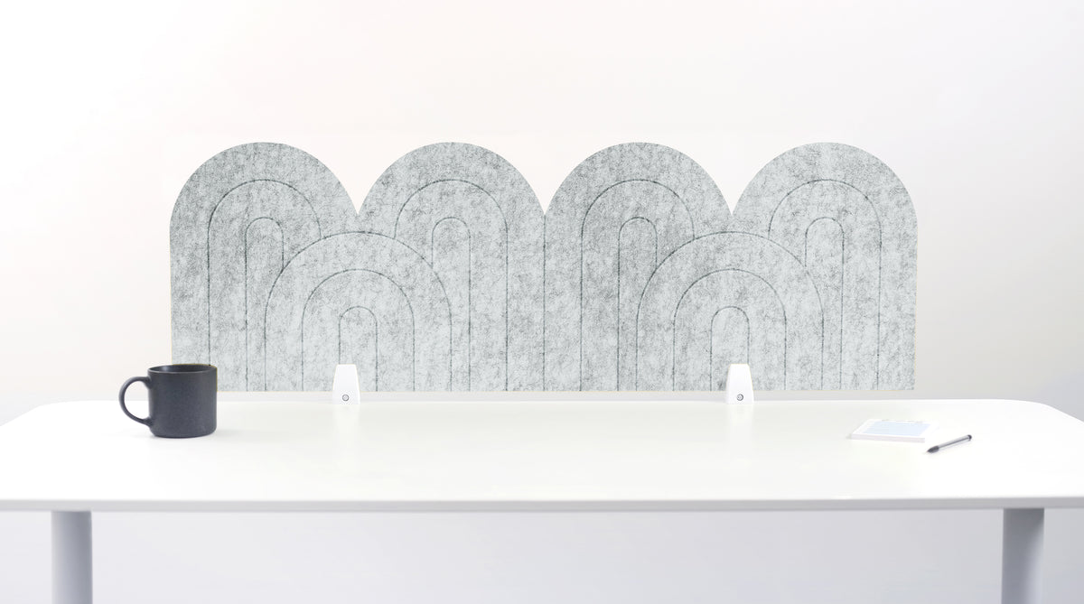 A nickel colored felt desk divider with a wavy, layered arch pattern, shown standing behind a white desk with a black mug on the left and a notepad on the right.