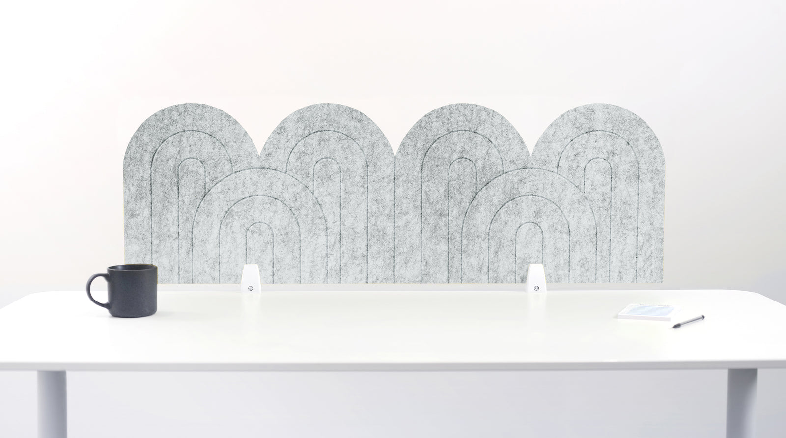 A nickel colored felt desk divider with a wavy, layered arch pattern, shown standing behind a white desk with a black mug on the left and a notepad on the right.