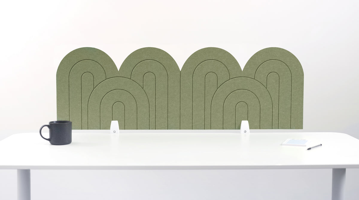 An olive felt desk divider with a wavy, layered arch pattern, shown standing behind a white desk with a black mug on the left and a notepad on the right.