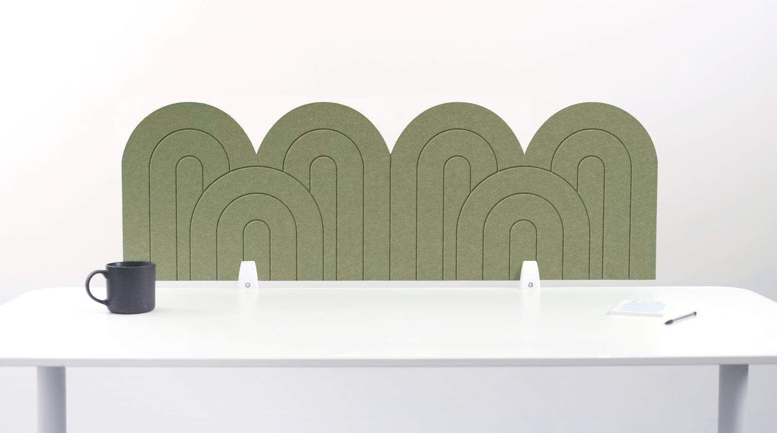 An olive felt desk divider with a wavy, layered arch pattern, shown standing behind a white desk with a black mug on the left and a notepad on the right.