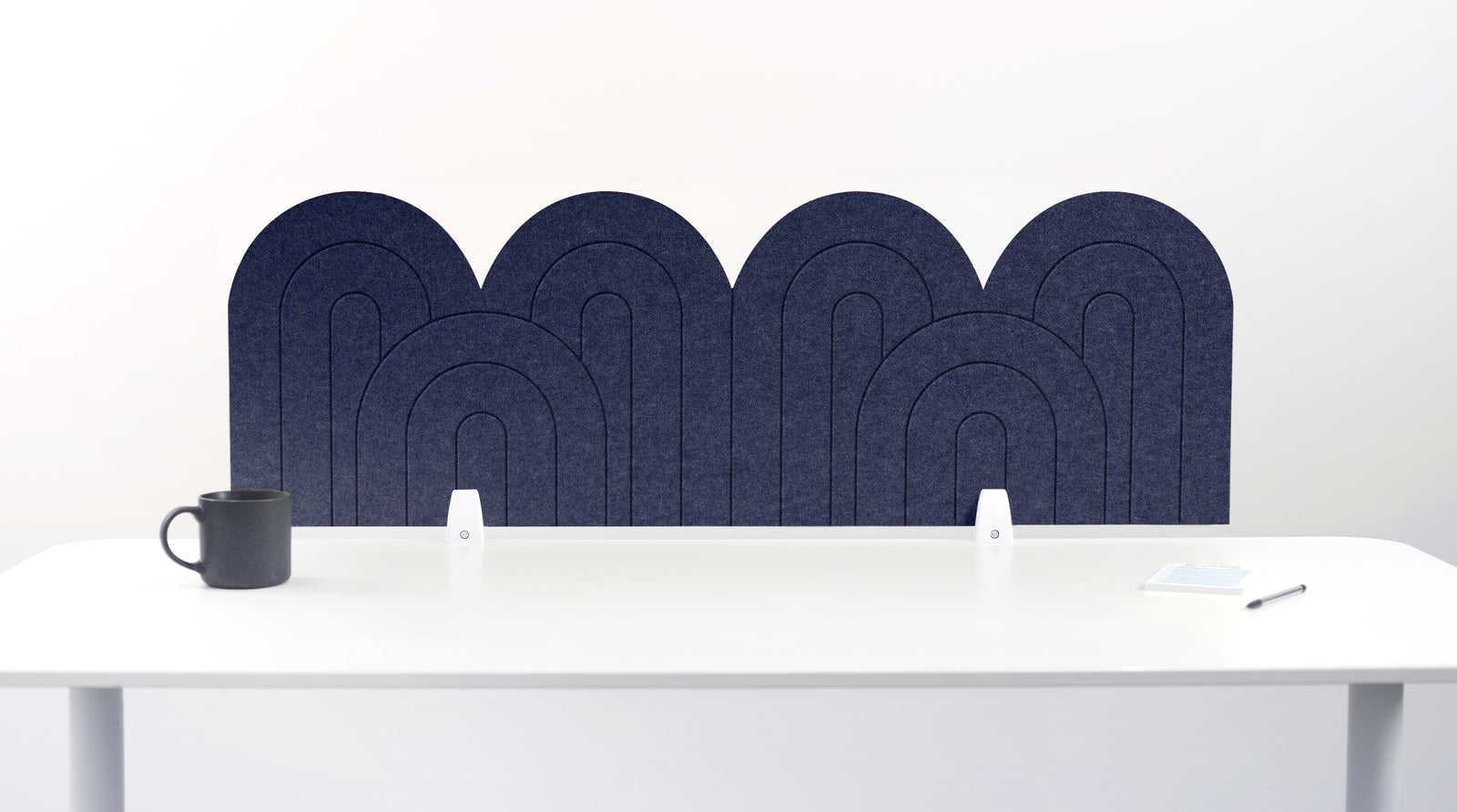 A navy blue felt desk divider with a wavy, layered arch pattern, shown standing behind a white desk with a black mug on the left and a notepad on the right.