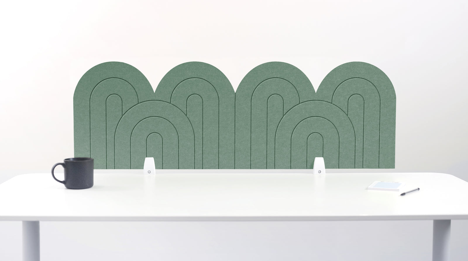 A long green palm felt desk divider with a wavy, layered arch pattern, shown standing behind a white desk with a black mug on the left and a notepad on the right.
