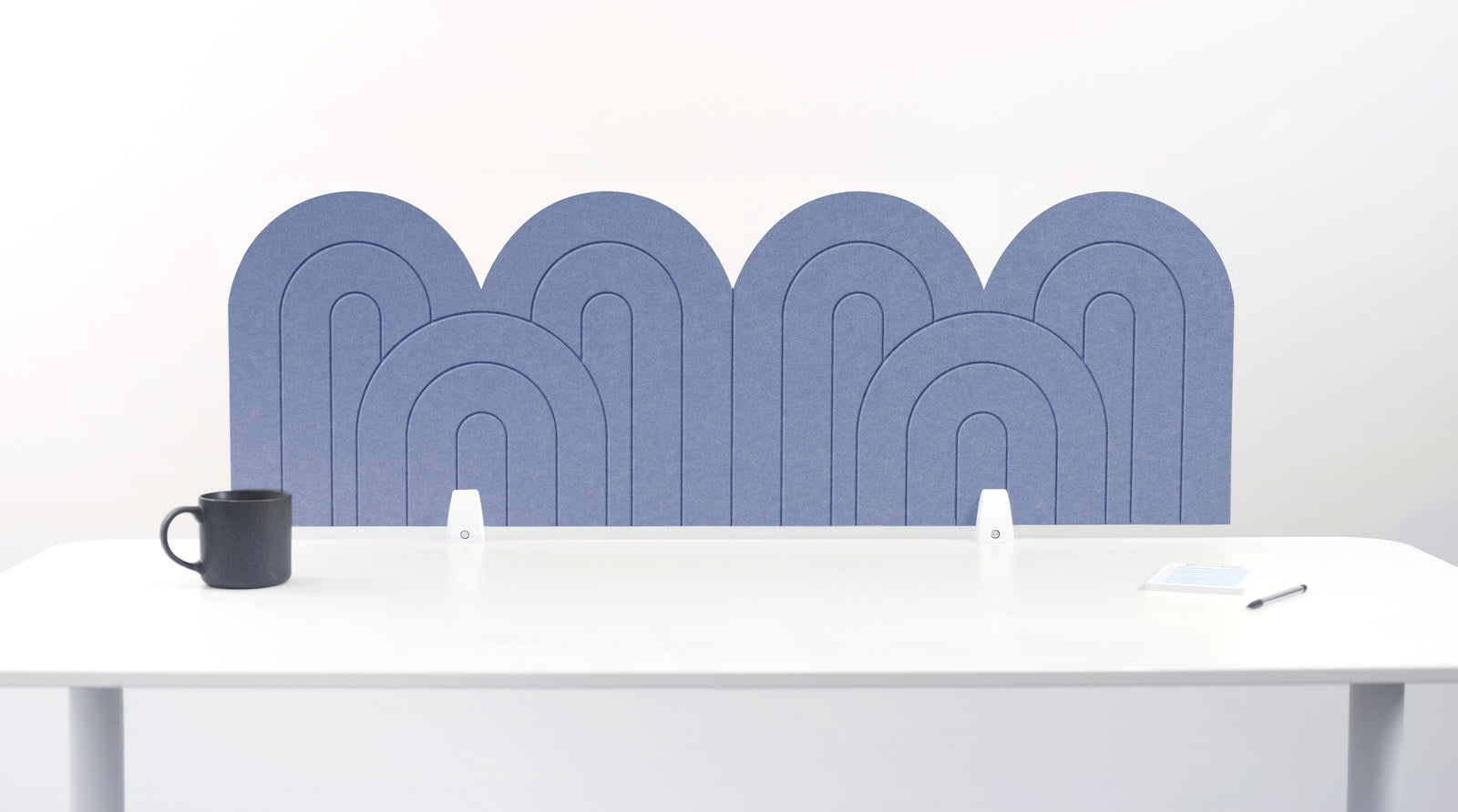 A periwinkle blue felt desk divider with a wavy, layered arch pattern, shown standing behind a white desk with a black mug on the left and a notepad on the right.