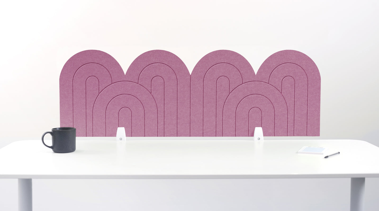 A pink felt desk divider with a wavy, layered arch pattern, shown standing behind a white desk with a black mug on the left and a notepad on the right.