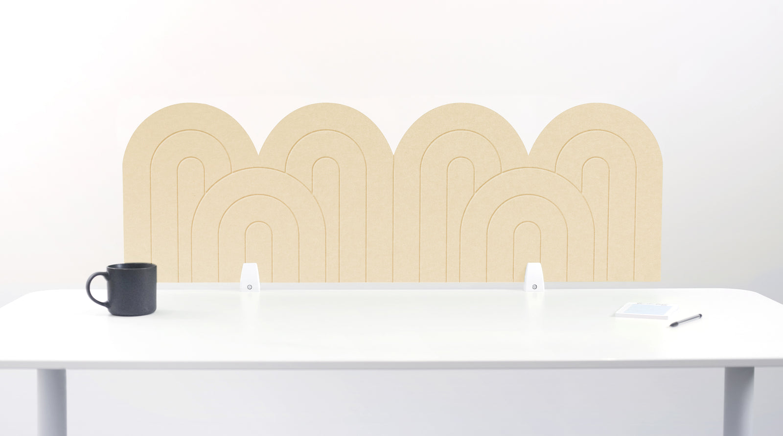 A yellow cream colored felt desk divider with a wavy, layered arch pattern, shown standing behind a white desk with a black mug on the left and a notepad on the right.