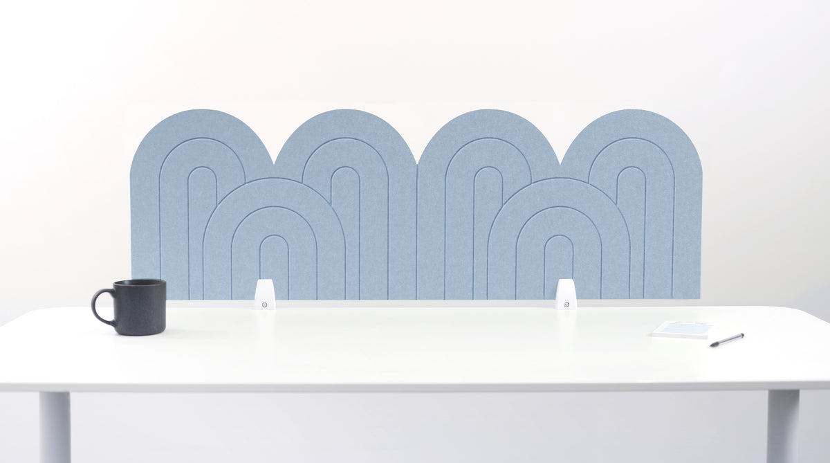A light blue felt desk divider with a wavy, layered arch pattern, shown standing behind a white desk with a black mug on the left and a notepad on the right.