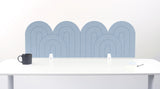 A light blue felt desk divider with a wavy, layered arch pattern, shown standing behind a white desk with a black mug on the left and a notepad on the right.