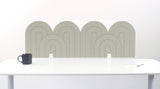 A sage green felt desk divider with a wavy, layered arch pattern, shown standing behind a white desk with a black mug on the left and a notepad on the right.