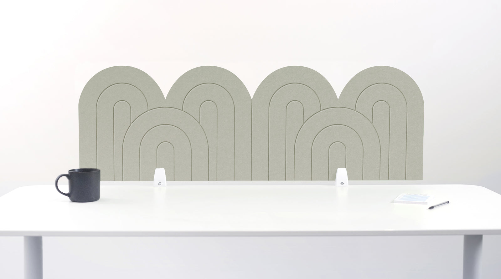 A sage green felt desk divider with a wavy, layered arch pattern, shown standing behind a white desk with a black mug on the left and a notepad on the right.