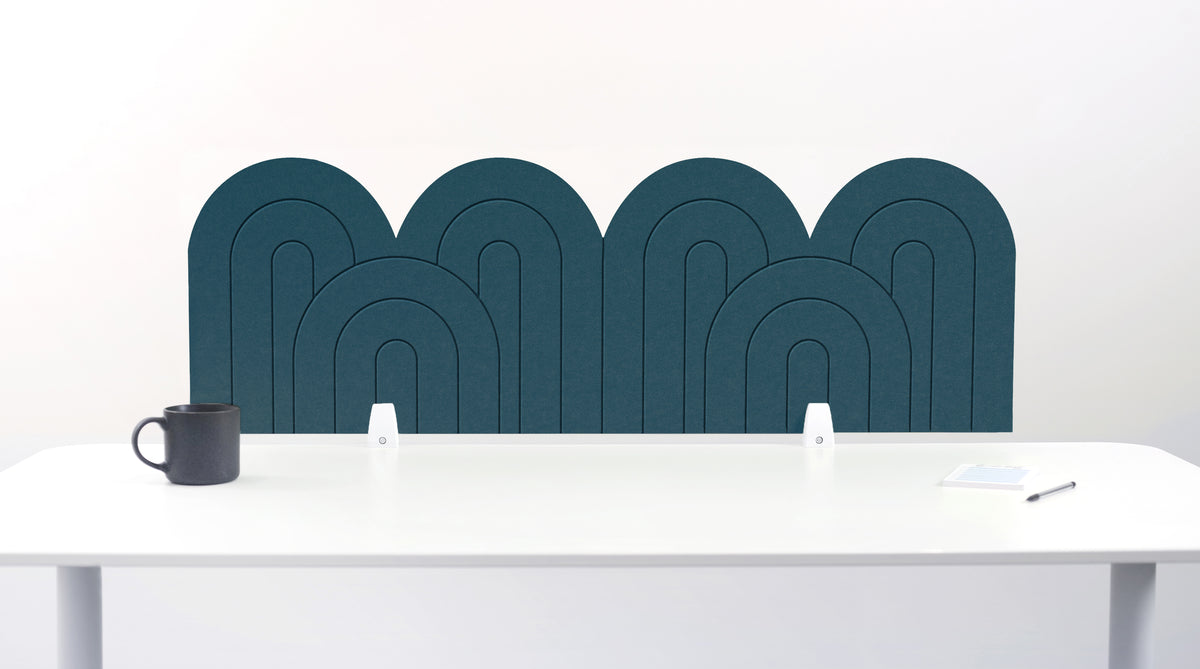 A slate blue felt desk divider with a wavy, layered arch pattern, shown standing behind a white desk with a black mug on the left and a notepad on the right.
