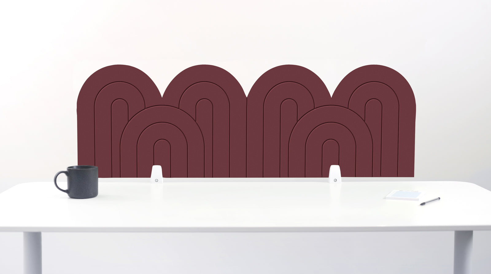 A brown felt desk divider with a wavy, layered arch pattern, shown standing behind a white desk with a black mug on the left and a notepad on the right.