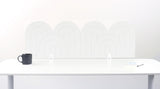 A white felt desk divider with a wavy, layered arch pattern, shown standing behind a white desk with a black mug on the left and a notepad on the right.