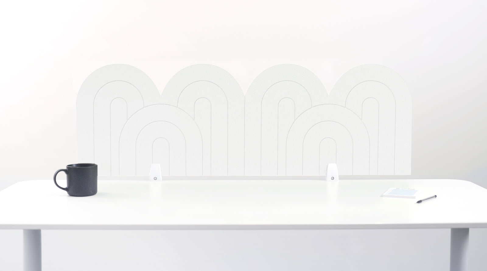 A white felt desk divider with a wavy, layered arch pattern, shown standing behind a white desk with a black mug on the left and a notepad on the right.