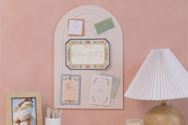 Decorative wall panel with framed pictures and notes on a pink wall, next to a lamp.