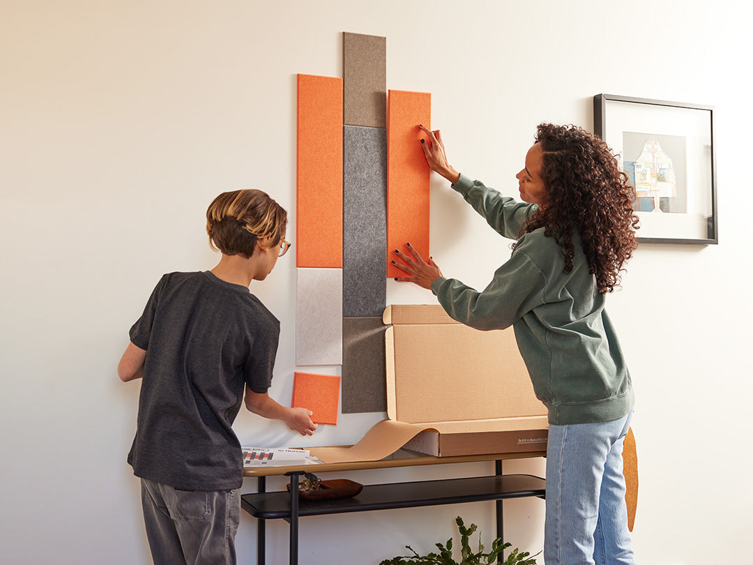 A Felt Right wall art design in orange, browns, and grey of felt that is pinnable on a wall above a side table with a mother and son setting it up
