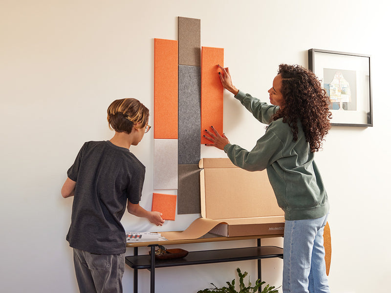 A Felt Right wall art design in orange, browns, and grey of felt that is pinnable on a wall above a side table with a mother and son setting it up