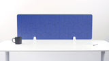 A blue acoustic desk divider with a scalloped wave pattern, mounted on a white desk with a black mug on the left and a small notebook on the right.