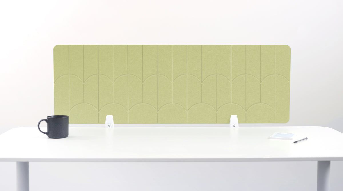 A light sage green felt desk divider with a scalloped wave pattern, mounted on a white desk with a black mug on the left and a small notepad on the right.