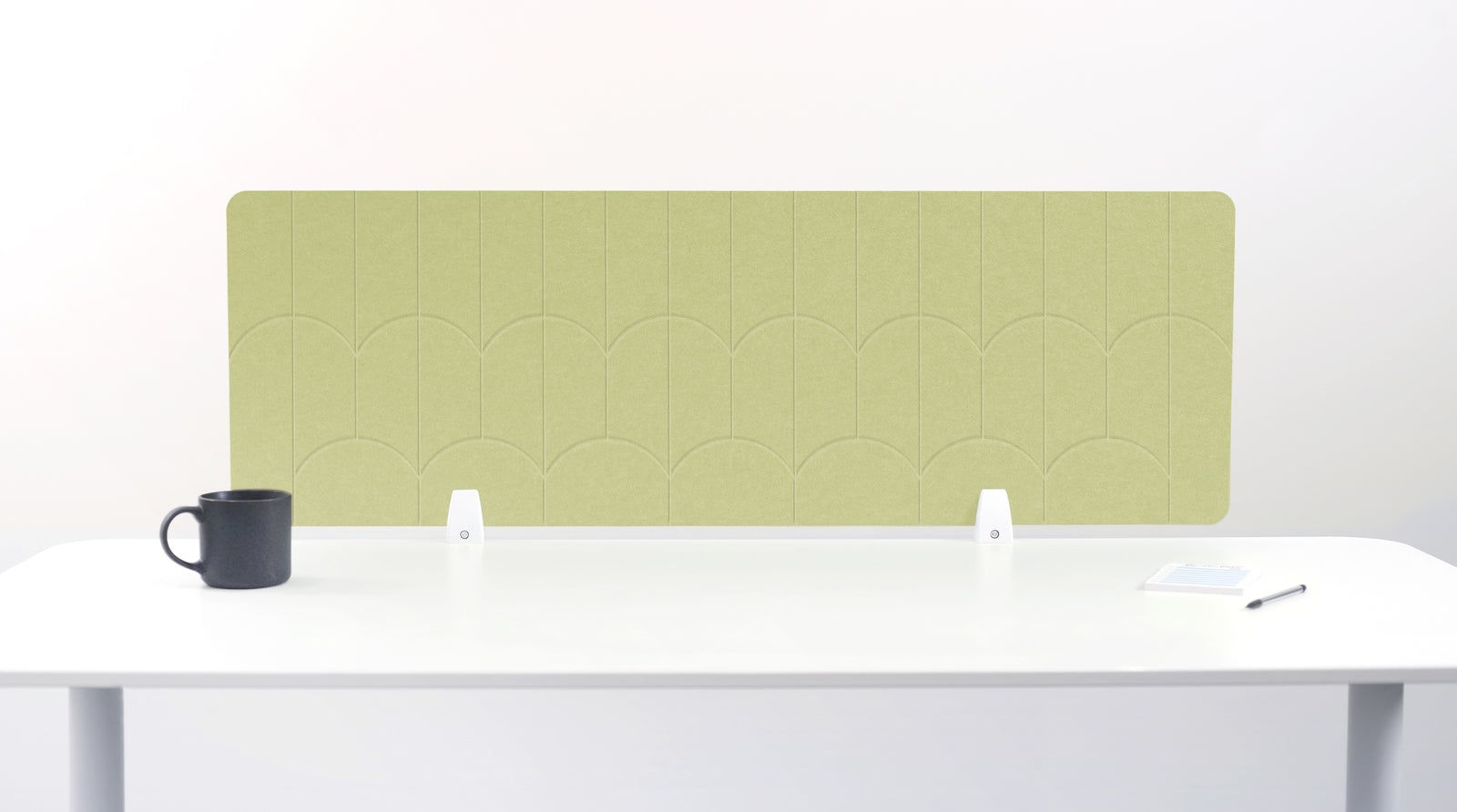 A light sage green felt desk divider with a scalloped wave pattern, mounted on a white desk with a black mug on the left and a small notepad on the right.