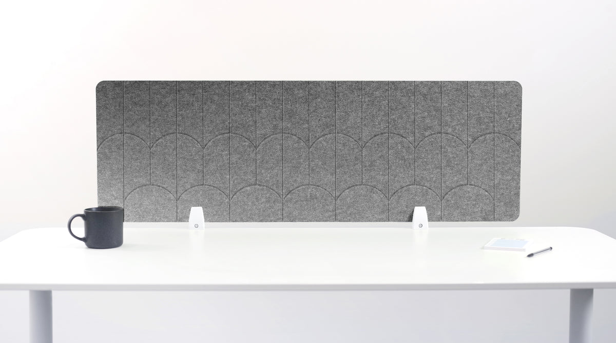 A light gray felt desk divider with a scalloped wave pattern, mounted on a white desk with a black mug on the left and a small notepad on the right.