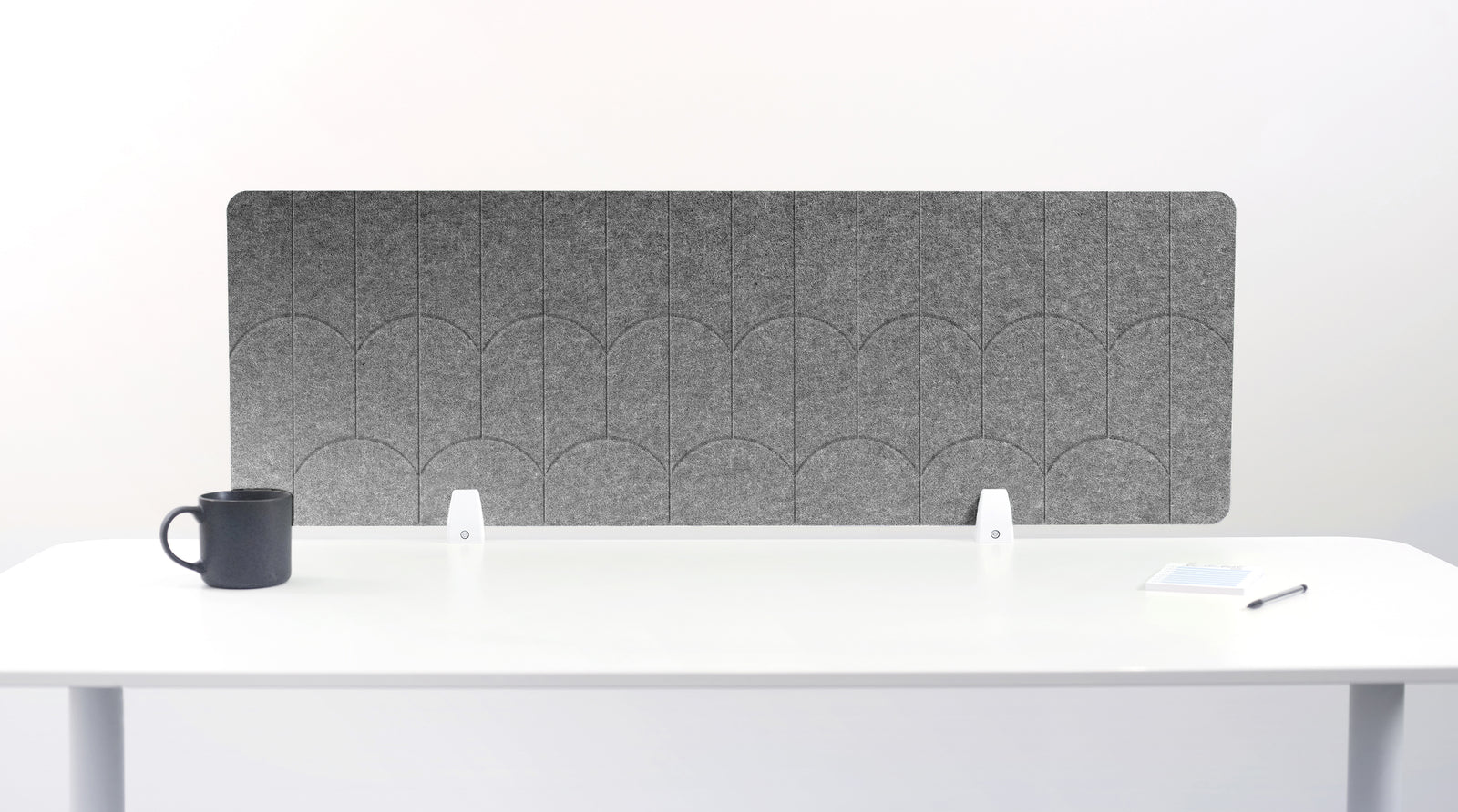 A light gray felt desk divider with a scalloped wave pattern, mounted on a white desk with a black mug on the left and a small notepad on the right.