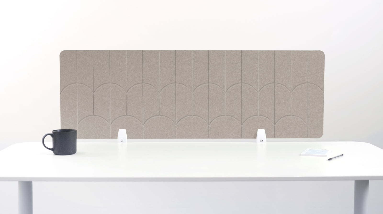 A light brown felt desk divider with a scalloped wave pattern, mounted on a white desk with a black mug on the left and a small notepad on the right.